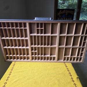 May include: A rectangular wooden printer's tray featuring numerous small, open compartments of varying sizes arranged in a grid pattern. The tray has a dark outer frame and a light-colored wood interior with a metal handle attached to the top edge.