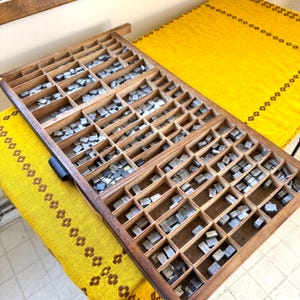 May include: A vintage wooden printer's tray filled with metal type pieces. The tray is divided into numerous small compartments, each holding individual letters and symbols. The wood has a warm, aged tone, and the metal type has a silver-gray color.