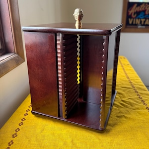 May include: A dark cherry-stained wooden CD storage cabinet with a decorative gold-colored finial on top. The cabinet has multiple slots for CD storage and is displayed on a yellow tablecloth.
