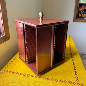 May include: A square, dark brown wooden media storage rack with four open sides featuring horizontal slots for organizing discs. The unit has a flat top surface with a small, decorative, metallic gold-colored finial knob centered on top.