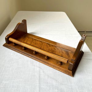 May include: A wooden wall shelf with a warm brown finish. The shelf features a long, flat surface with a rounded bar and several small pegs below. The design includes two upright supports with decorative cutouts.