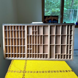 May include: A rectangular wooden printer's tray featuring numerous small, open compartments of varying sizes arranged in a grid pattern. The tray has a light brown wood finish with a dark frame and a flat, light-colored backing in each section.