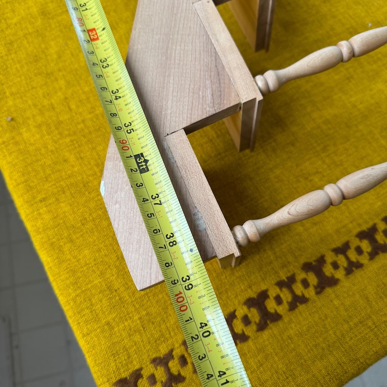 May include: A light-colored wooden shelf with two tiers and turned legs. A yellow measuring tape shows the shelf is approximately 38 inches long. The shelf is on a yellow and brown patterned surface.