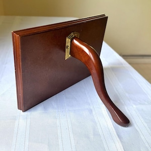 May include: A rectangular, dark brown wooden picture frame with a curved, wooden stand. The stand is attached to the back of the frame with a brass-colored metal bracket. The frame is displayed on a white surface.
