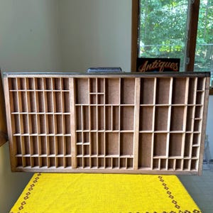 May include: A rectangular wooden printer's drawer featuring a grid of numerous small, open compartments of varying sizes. The frame is dark brown, and the interior dividers are light tan, creating a structured storage display case.