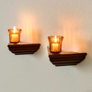 May include: Two wall-mounted candle holders with lit candles. Each holder has a dark brown, tiered shelf design. The candles are in amber-colored glass votives, casting a warm glow. The holders are mounted on a white wall.