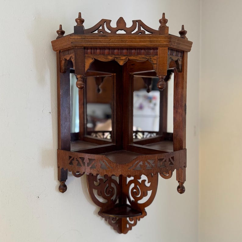 May include: A brown wooden corner shelf featuring two mirrored back panels, a flat top surface, and a lower triangular shelf. The structure is adorned with intricate cut-out scrollwork patterns along the bottom and top edges, with small finials.