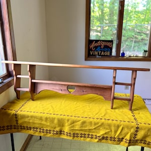 May include: A long, wooden shelf with a natural finish, featuring a heart-shaped cutout and two small shelves on either side. The shelf is displayed on a yellow tablecloth with a brown patterned border. The background includes a window and a sign that reads "Antiques & Vintage."