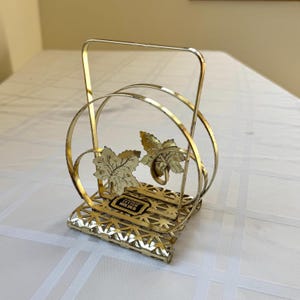 May include: A gold-toned metal letter holder with a decorative leaf design. The holder features a rectangular base with a geometric pattern, a curved central section, and a handle. The leaves are detailed with veins and add a touch of nature.