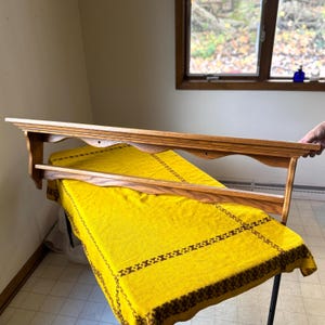 Puede incluir: Una estantería de madera marrón claro con un borde frontal festoneado y una estantería inferior. La estantería se muestra sobre un mantel amarillo. La estantería es probablemente de madera.