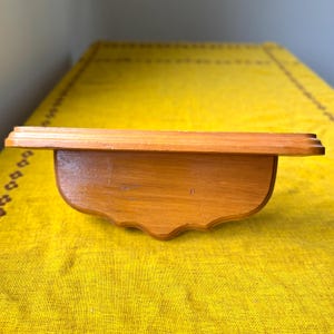 May include: A small, wooden shelf with a curved bottom and a decorative edge. The shelf is made of light brown wood and is set against a yellow background.