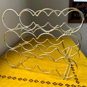 May include: A gold-colored metal wine rack with a three-tier design. Each tier has a wavy pattern with circular openings to hold wine bottles. The rack is displayed on a yellow patterned surface.