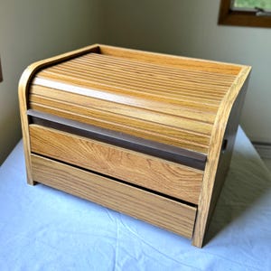 May include: A wooden bread box with a roll-top lid and two drawers. The box is made of light-colored wood with a natural grain. The roll-top lid has horizontal grooves, and the drawers have a simple, clean design. The box is rectangular with rounded edges.