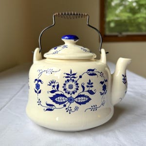 May include: Cream-colored ceramic teapot with blue floral designs. Features a metal handle with a wooden grip and a matching lid. The teapot has a spout and is decorated with blue flowers and leaves.