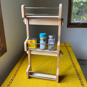 May include: A wooden spice rack with three shelves and metal bars. The rack holds a yellow mustard tin, a blue-topped garlic container, and a jar of oregano. The rack is on a yellow tablecloth.
