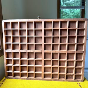 May include: A rectangular wooden printer's tray featuring a grid of numerous small, square compartments. The organiser is constructed from light brown wood with a natural grain finish and includes a single small metal knob at the top centre.