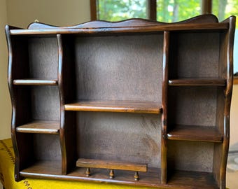 May include: A dark brown wooden wall shelf with multiple compartments and shelves. The shelf has a wavy top edge and a small peg rail at the bottom. The wood grain is visible, and the shelf appears to be vintage.