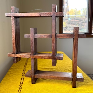May include: A handcrafted wooden shelf with a geometric design. The shelf is constructed from dark-stained wood and features multiple horizontal and vertical supports, creating a multi-tiered display space. The wood grain is visible, adding to the rustic aesthetic.