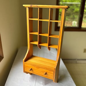 May include: A tall, wooden wall shelf with a natural finish. It features multiple small cubby shelves and a drawer with two wooden knobs. The shelf has a unique, curved design on the sides and is suitable for displaying small items.