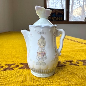 May include: A white ceramic paprika jar with a lid and handle. The jar features a cartoon illustration of a girl holding a basket of grapes, with the word "Paprika" written above. The lid has a decorative butterfly-shaped handle.