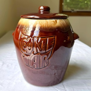 May include: A brown ceramic cookie jar with a lid. The jar has a glossy finish and the word "COOKIE JAR" is embossed on the front. The jar has a two-tone color scheme, with a lighter brown at the top and a darker brown at the bottom.