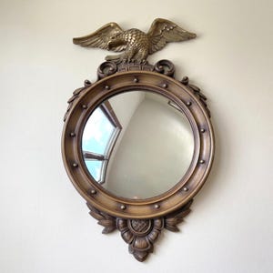 Puede incluir: Un espejo redondo de estilo antiguo con un marco de color bronce y un emblema de águila en la parte superior. El espejo tiene un borde decorativo con remaches y detalles florales en la parte inferior. El águila está posada sobre el espejo, con las alas extendidas.