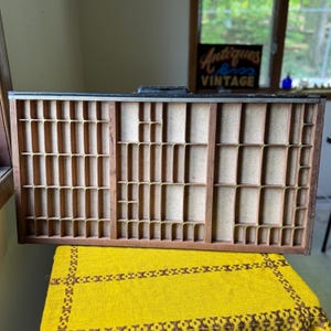 May include: A vintage wooden printer's tray with numerous small compartments. The tray is made of dark wood and has a rectangular shape. The compartments are separated by thin wooden dividers. The tray sits on a yellow patterned cloth.