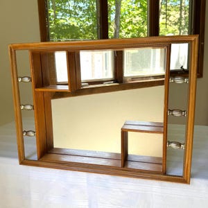 May include: A rectangular wooden framed mirror with built-in shelves. The mirror has a light brown finish with decorative silver-toned handles on the sides. The shelves are positioned within the frame, creating a unique storage space.