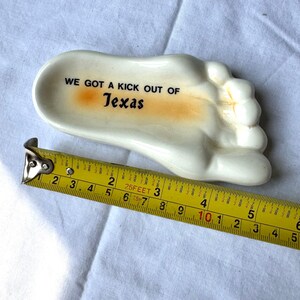 May include: A ceramic foot-shaped dish with the text "WE GOT A KICK OUT OF Texas" in black. The dish is cream-colored with an orange accent. The dish measures approximately 5 inches in length.