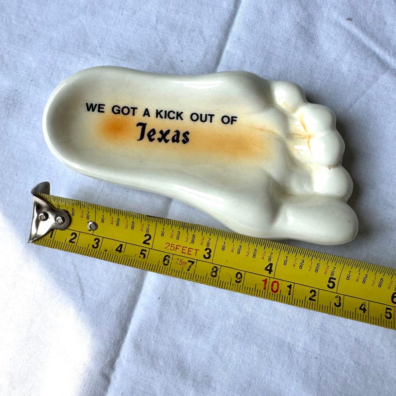 May include: A ceramic foot-shaped dish with the text "WE GOT A KICK OUT OF Texas" in black. The dish is cream-colored with an orange accent. The dish measures approximately 5 inches in length.