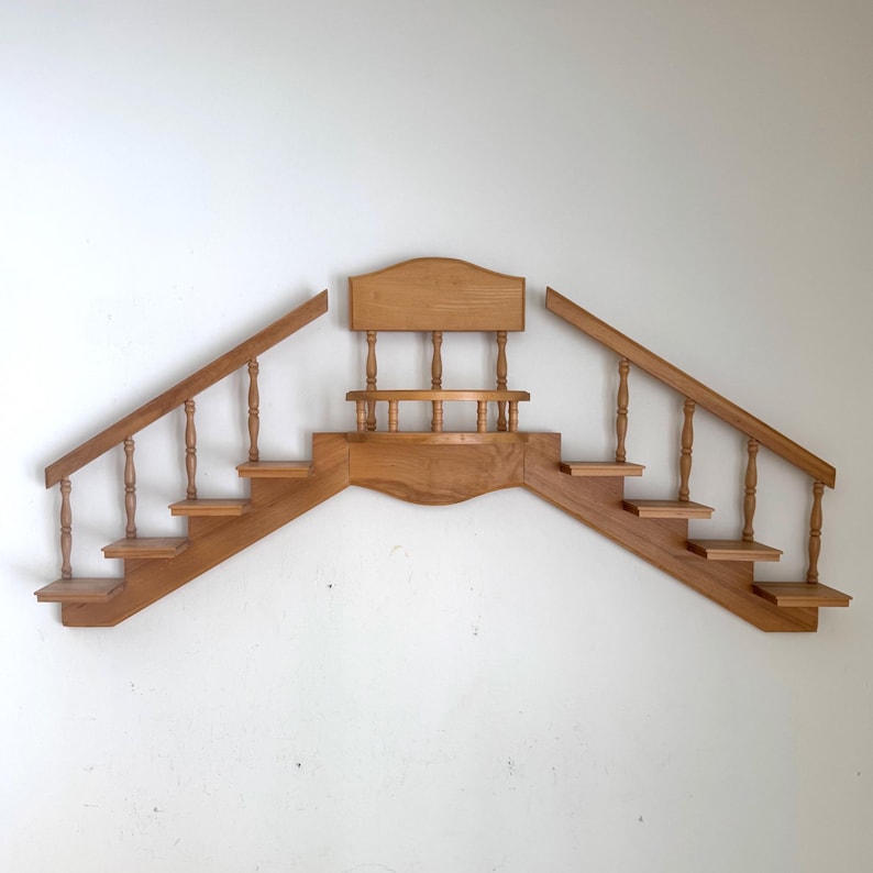 May include: Wooden wall shelf in the shape of a staircase with a central platform. The shelf has two staircases with balustrades and a small platform in the middle. The wood is a light brown color.