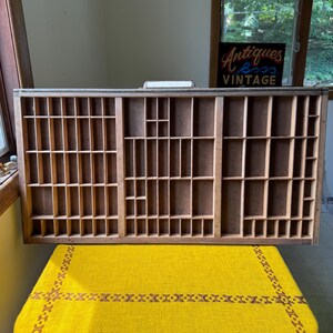 May include: A rectangular wooden printer's tray featuring numerous small, open compartments of varying sizes arranged in a grid pattern. The tray is constructed from light brown wood with a flat, dark brown backing and a metal handle at the top center.