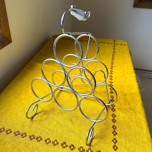May include: A silver-colored wine rack with a unique design. The rack holds multiple bottles and features a decorative handle. The rack is made of metal and has a circular design. The rack is on a yellow tablecloth.