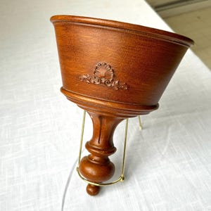 May include: A brown wooden planter with a decorative shell carving. The planter is supported by a gold-colored metal stand. The planter has a flared shape and a detailed, carved design.