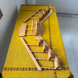 May include: A disassembled wooden dollhouse staircase set. The set includes multiple stair sections, railings, and a landing. The wood has a natural finish, and the pieces are arranged on a yellow patterned tablecloth.