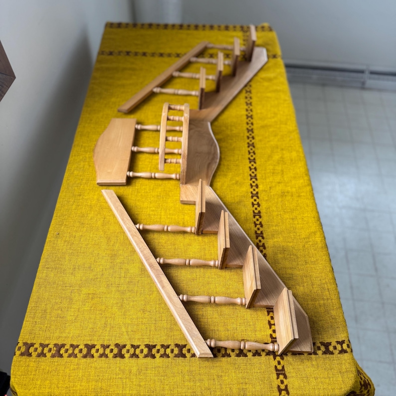 May include: A disassembled wooden dollhouse staircase set. The set includes multiple stair sections, railings, and a landing. The wood has a natural finish, and the pieces are arranged on a yellow patterned tablecloth.