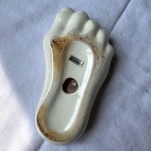 May include: A white ceramic foot-shaped ashtray with a brown-stained rim and a small hole. The bottom of the ashtray has a sticker that reads "MADE IN TAIWAN".