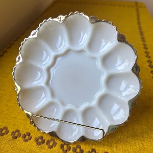 May include: A white, scalloped egg plate with a silver-colored trim. The plate has 12 egg indentations and a central well. The plate is displayed on a gold-colored metal stand, set against a yellow background.