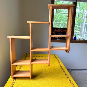 May include: A wooden, tiered shelf unit with a natural finish. The shelf has multiple rectangular platforms at varying heights, creating a stepped design. The unit is freestanding and appears to be made of solid wood.