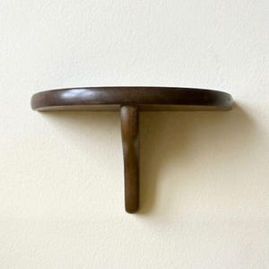 May include: A semi-circular wall-mounted shelf crafted from dark brown wood. The shelf features a smooth, curved top surface and a single vertical support bracket extending downward from the center, creating a simple, functional design.