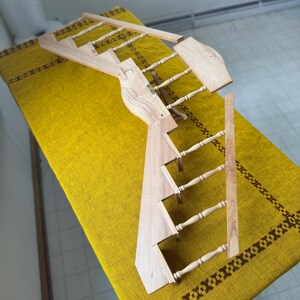 May include: A wooden wall-mounted display rack with a ladder-like design. The rack is made of light-colored wood and features multiple rungs for hanging items. The design includes a decorative element at the top and is placed on a yellow tablecloth.