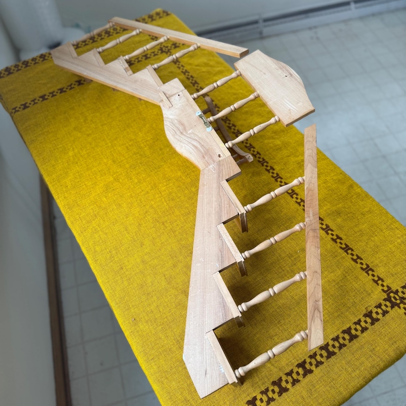 May include: A wooden wall-mounted display rack with a ladder-like design. The rack is made of light-colored wood and features multiple rungs for hanging items. The design includes a decorative element at the top and is placed on a yellow tablecloth.