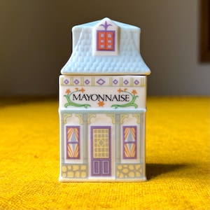 May include: A ceramic container shaped like a small house with a light blue roof and a white base. The front features the word MAYONNAISE in black text, surrounded by green vines and orange stars. It has purple windows and a central purple door.