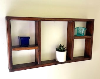 May include: A dark brown wooden wall shelf with four compartments. The shelf holds a small blue box, a white potted succulent, and a turquoise glass bowl. The shelf is mounted on a light-colored wall.