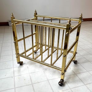 May include: A vintage brass magazine rack on wheels. The rack has a rectangular shape with vertical bars and decorative finials on the corners. The brass has a polished finish, and the wheels are small and ornate.