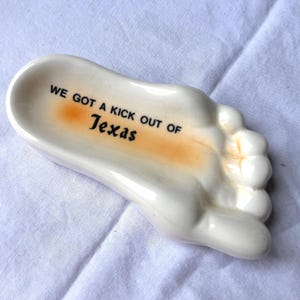 May include: A white ceramic foot-shaped dish with the text "WE GOT A KICK OUT OF Texas" in black and orange. The dish has a small, shallow bowl area and is resting on a white surface.