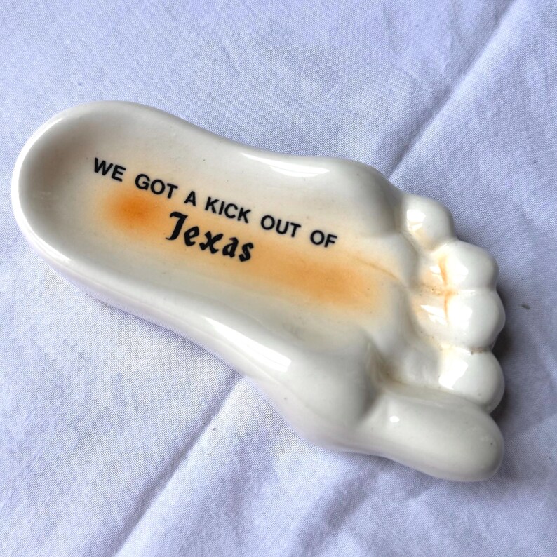 May include: A white ceramic foot-shaped dish with the text "WE GOT A KICK OUT OF Texas" in black and orange. The dish has a small, shallow bowl area and is resting on a white surface.
