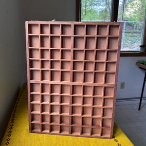 Könnte beinhalten: Ein Vintage-Druckertablett aus Holz mit zahlreichen kleinen, quadratischen Fächern. Das Tablett besteht aus rotbraunem Holz und ist für kleine Gegenstände oder Sammlerstücke konzipiert. Die gitterartige Struktur ist ideal zum Organisieren und Präsentieren verschiedener Objekte.