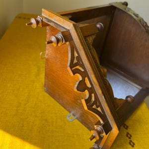 May include: A brown wooden wall shelf featuring intricate cut-out scrollwork along the side supports and decorative finials at the corners. The structure includes a flat top surface and a back panel with metal mounting hardware attached.
