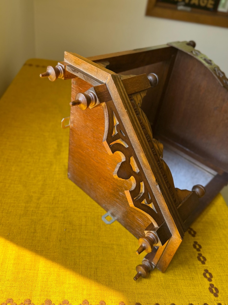 May include: A brown wooden wall shelf featuring intricate cut-out scrollwork along the side supports and decorative finials at the corners. The structure includes a flat top surface and a back panel with metal mounting hardware attached.